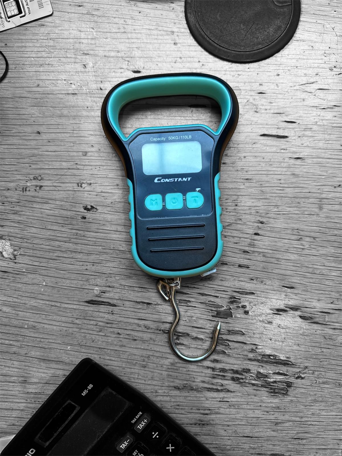 Constant electronic hanging luggage scale on a wooden desk beside a calculator