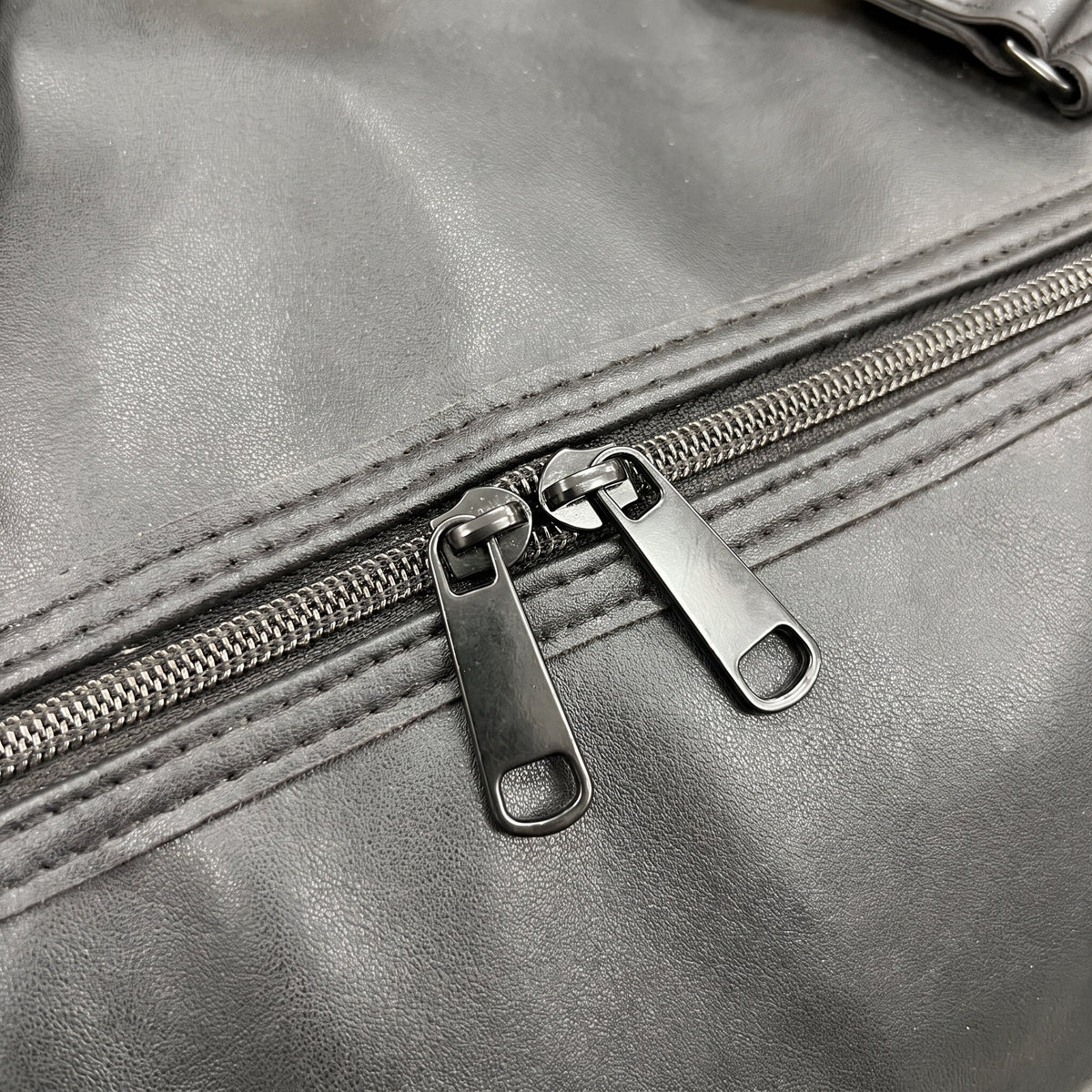 Close up of the faux leather grain and the twin zip sliders on the JUSTPACK Belgrave wheeled holdall in black faux leather
